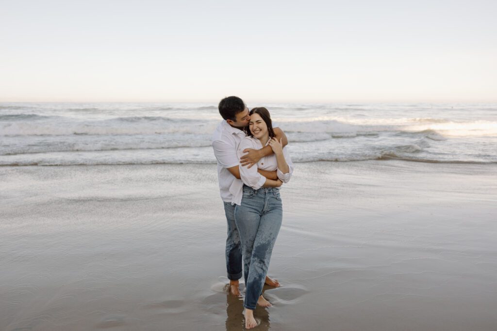 A couple cozies up on the beach wearing matching outfits of white tops and blue jeans.