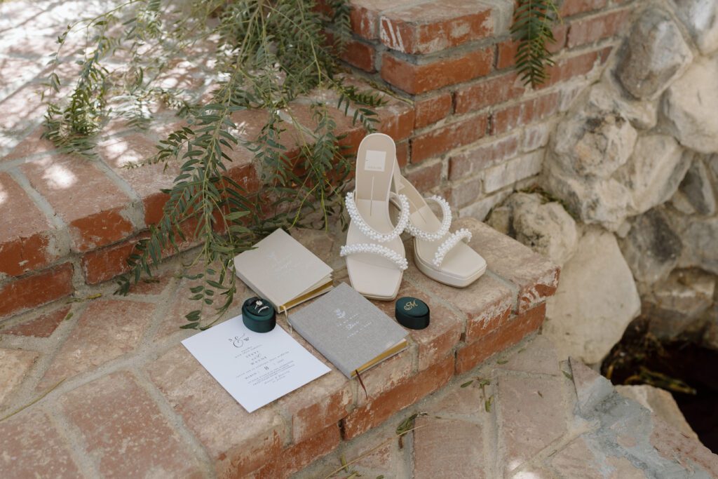 Simple wedding flat lay of bridal heels, ring boxes, vow books, and a wedding invitation 