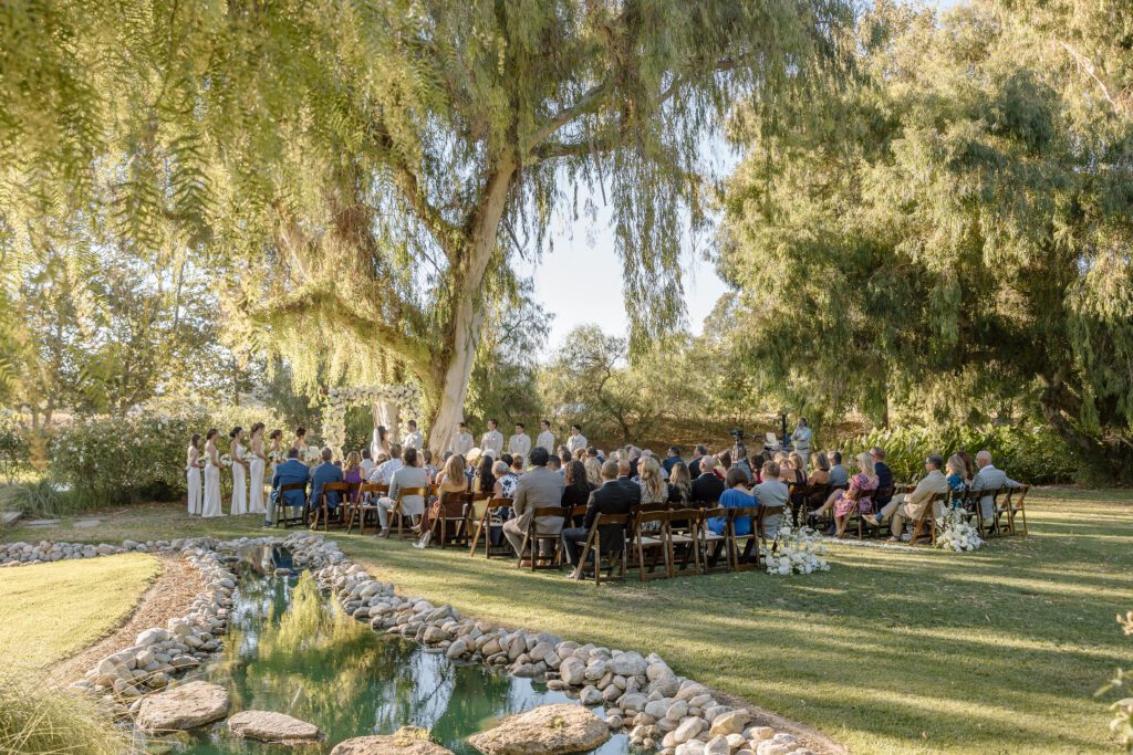 On a warm summer's day, guests sit beneath a canopy of willow trees next to a babbling brook as a bride and groom exchange heartfelt vows during their Southern California wedding.