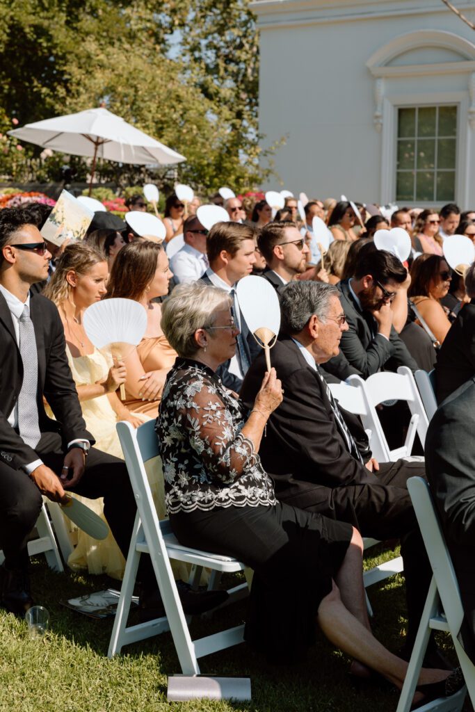 Guests fan themselves with paper hand-fans during a Southern California outdoor wedding ceremony, photographed by Laura Burns Photography.