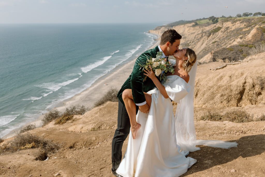 Groom dips bride in a romantic kiss on a coastal cliff during a beautiful Southern California beach elopement, with ocean views and rugged shoreline in the background.