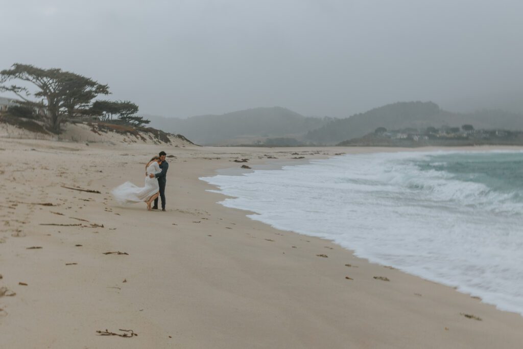 Intimate Southern California beach elopement with bride and groom embracing on a foggy coastline surrounded by dramatic ocean waves and misty hills.