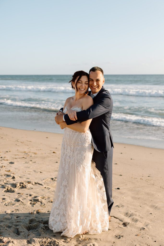 A groom lovingly wraps his arms around his bride on the beach following their tropical Southern California outdoor wedding, photographed by Laura Burns Photography.