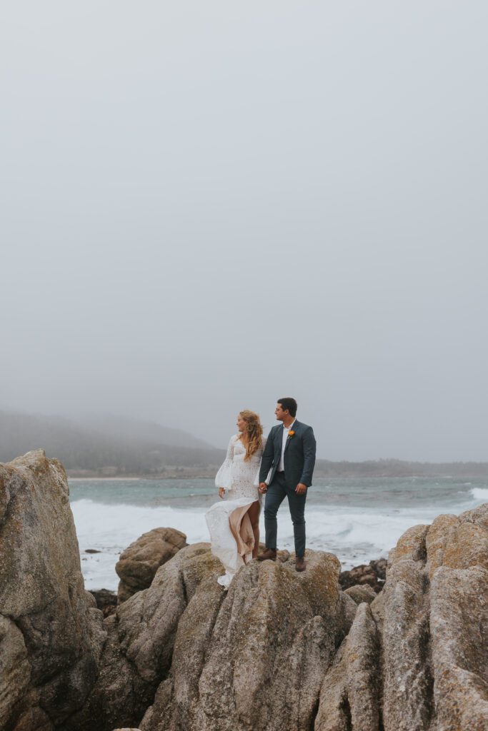 Bride and groom standing on top of a rock at the beach with a cloudy stormy sky behind them.